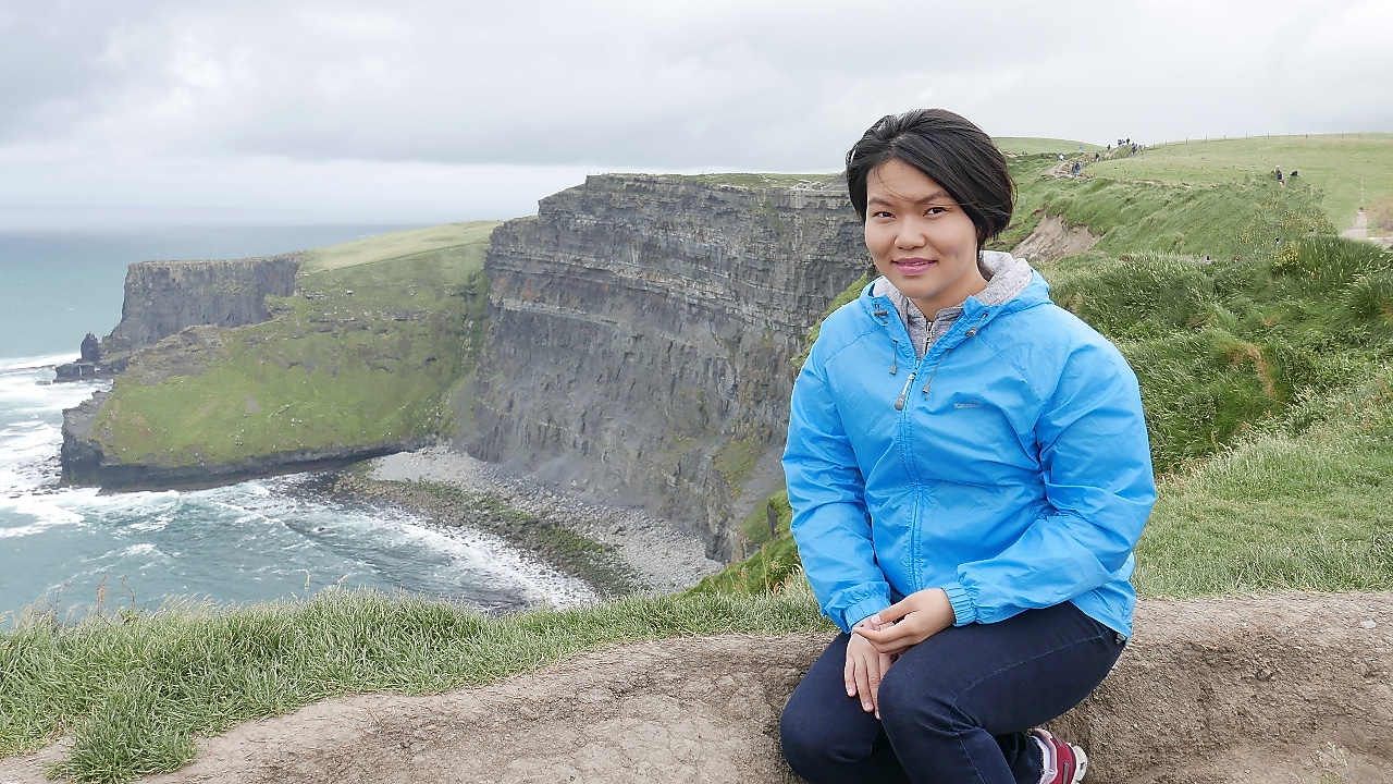 A short hair lady in a blue jacket posing on top of a mountain with sea background