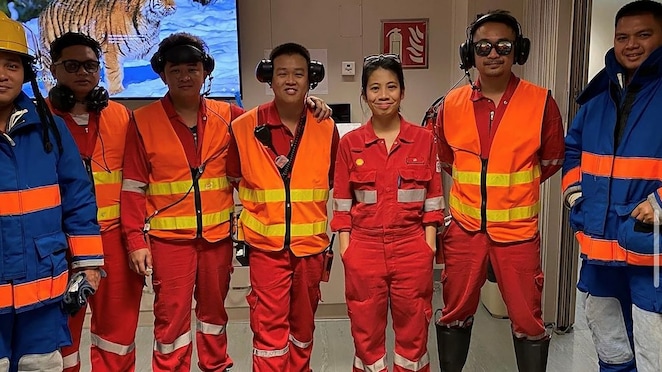 Group of offshore workers in safety gear posing indoors, wearing coveralls, high-visibility vests, hearing protection and boots
