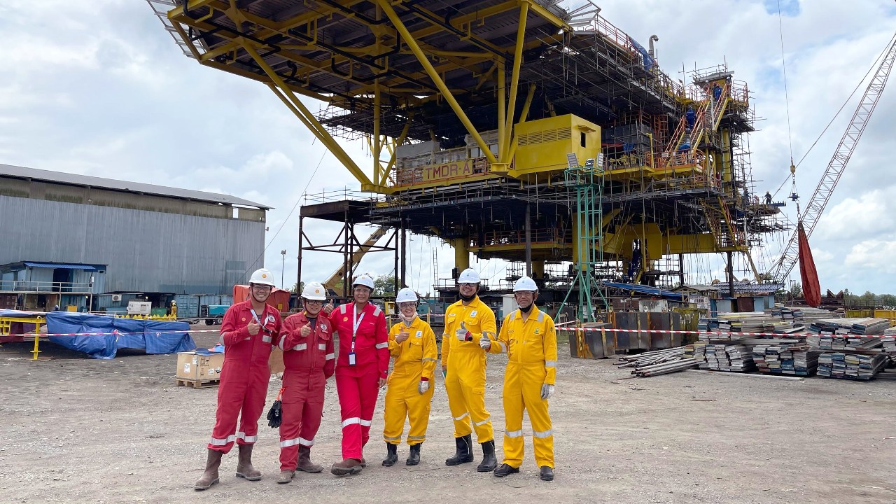 A group of engineers wearing coverall and white hard hat posing thumbs up and smiling at an onshore area