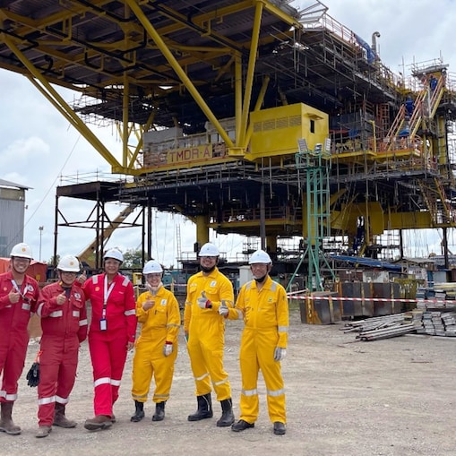 A group of engineers wearing coverall and white hard hat posing thumbs up and smiling at an onshore area