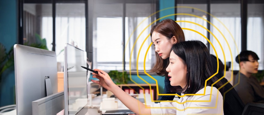 Two people working together at a modern office desk, focusing on a computer monitor. One person is pointing at the screen with a pen while the other observes. The desk has organized stationery items, documents, and a keyboard. Large windows in the background allow natural light into the room, and there are plants and shelves visible behind them.