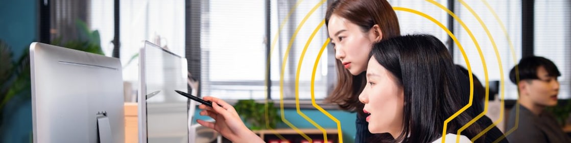 Two people working together at a modern office desk, focusing on a computer monitor. One person is pointing at the screen with a pen while the other observes. The desk has organized stationery items, documents, and a keyboard. Large windows in the background allow natural light into the room, and there are plants and shelves visible behind them.