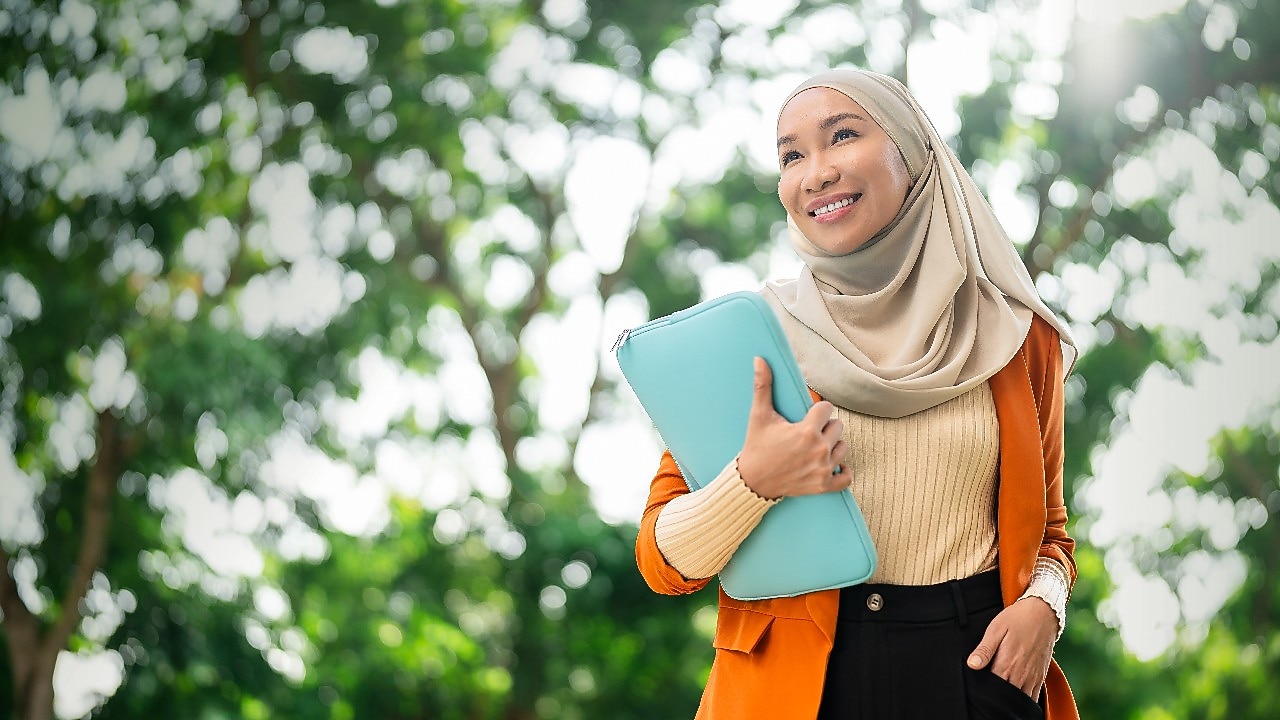 A person wearing a beige headscarf, a light-colored ribbed top, and an orange blazer is standing outdoors in a green park setting. The individual is holding a light blue laptop or folder against the chest with one hand. The background features lush green trees with sunlight filtering through the leaves, creating a bright and natural atmosphere. 