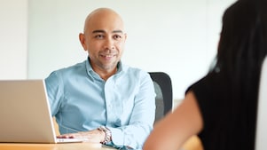 A colleague seated in front of his laptop and listening to his colleague across the table.