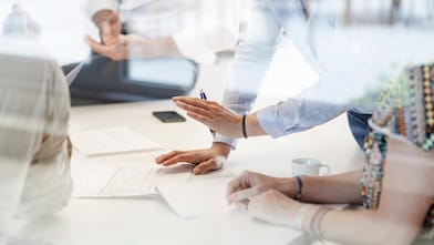 A team where just their hands are seen in the image, are having a planning meeting with papers on the table to refer to.