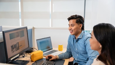 Two individuals are seated at a workstation in an office setting. One person is using a desktop computer with a large monitor displaying a webpage, while the other is sitting beside them. A laptop is placed next to the monitor, along with a yellow pouch and a yellow mug on the desk. The workspace has partition panels and a bright, well-lit background with large windows.