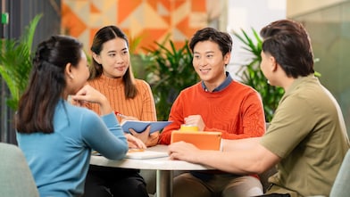 Four people are seated around a round table in a modern office setting, engaged in a discussion. Two individuals are holding notebooks, and a bright yellow mug is placed on the table. The background features indoor plants and a wall with geometric orange patterns, creating a vibrant and collaborative atmosphere. 