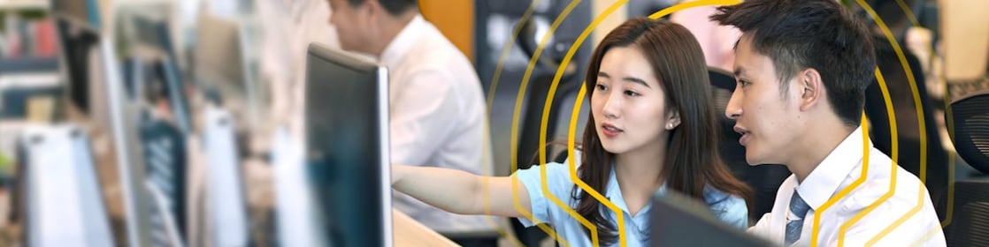 Two people sitting side by side at a desk in an open office environment, looking at a computer monitor. One person is pointing at the screen while the other observes. Multiple computer monitors and office supplies are visible on the desks, with other colleagues working in the background. The setting includes bright lighting, plants, and a modern workspace design.