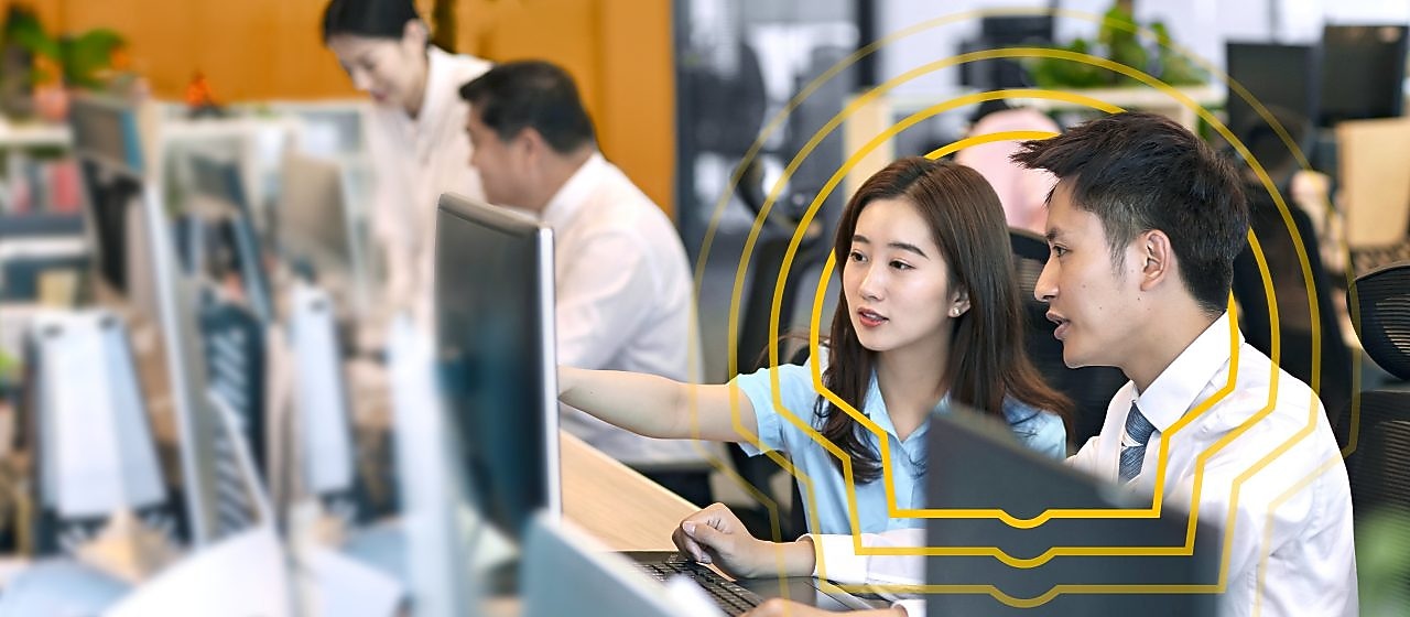 Two people sitting side by side at a desk in an open office environment, looking at a computer monitor. One person is pointing at the screen while the other observes. Multiple computer monitors and office supplies are visible on the desks, with other colleagues working in the background. The setting includes bright lighting, plants, and a modern workspace design.