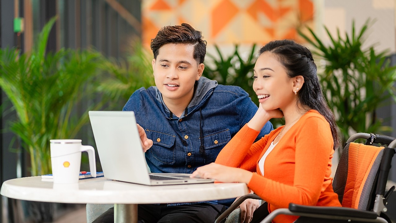 Two individuals are seated at a round table in a modern indoor setting, working together on a laptop. One person is wearing an orange cardigan and is seated in a wheelchair, while the other is wearing a dark blue denim shirt and pointing at the laptop screen. A white mug with a logo is placed on the table, along with a notebook and pen. The background features green plants and a wall with geometric orange patterns, creating a bright and collaborative atmosphere.