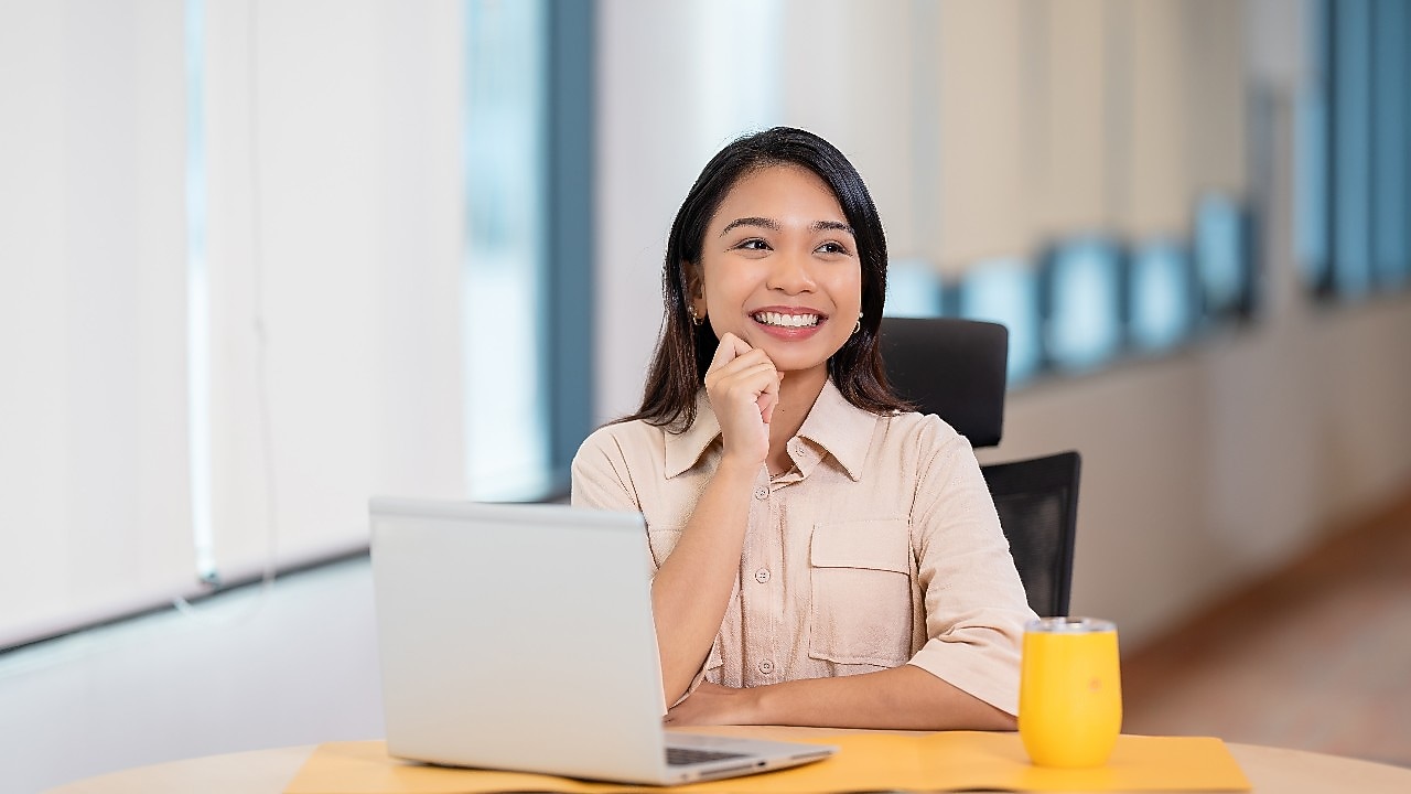A person is seated at a desk in a modern office setting with large windows in the background. On the desk, there is an open silver laptop and a bright yellow mug placed on a yellow mat. The person is wearing a light beige button-up shirt and is resting one hand near the chin while looking toward the laptop. The environment appears well-lit with natural light.