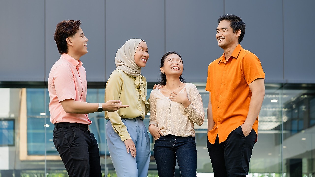 Four individuals are standing outdoors in front of a modern glass building, engaged in conversation. They are dressed in casual attire, including light-colored tops, an orange shirt, and a beige headscarf. The setting features a contemporary architectural backdrop with large windows and a gray panel wall.​