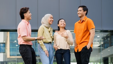 Four individuals are standing outdoors in front of a modern glass building, engaged in conversation. They are dressed in casual attire, including light-colored tops, an orange shirt, and a beige headscarf. The setting features a contemporary architectural backdrop with large windows and a gray panel wall.​