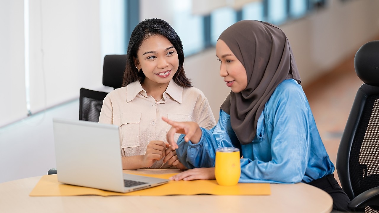 Two individuals are seated at a round table in a modern office setting. They are engaged in a discussion while looking at an open laptop placed on the table. One person is wearing a light-colored shirt, and the other is wearing a blue long-sleeve top with a headscarf. A bright yellow mug is also on the table, along with a yellow desk mat. The background shows large windows with natural light coming in.​  ​