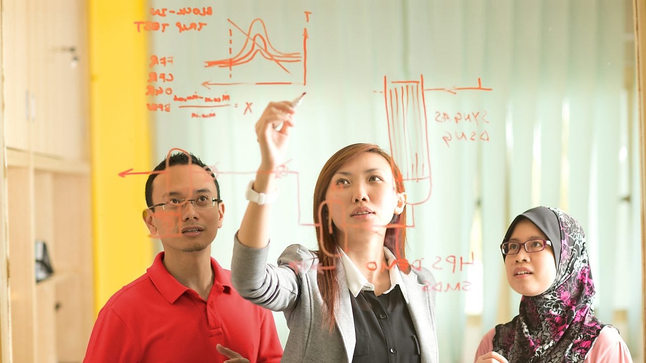 Colleagues gathered standing and discussing metrics on a glass board