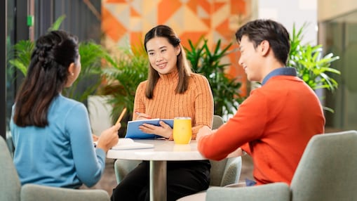 Three people are seated around a small round table in a modern office setting, engaged in a discussion. One person is holding a blue notebook, another has a yellow mug on the table, and the third appears to be gesturing while speaking. The background features green indoor plants and a wall with geometric orange patterns, creating a bright and collaborative atmosphere.​
