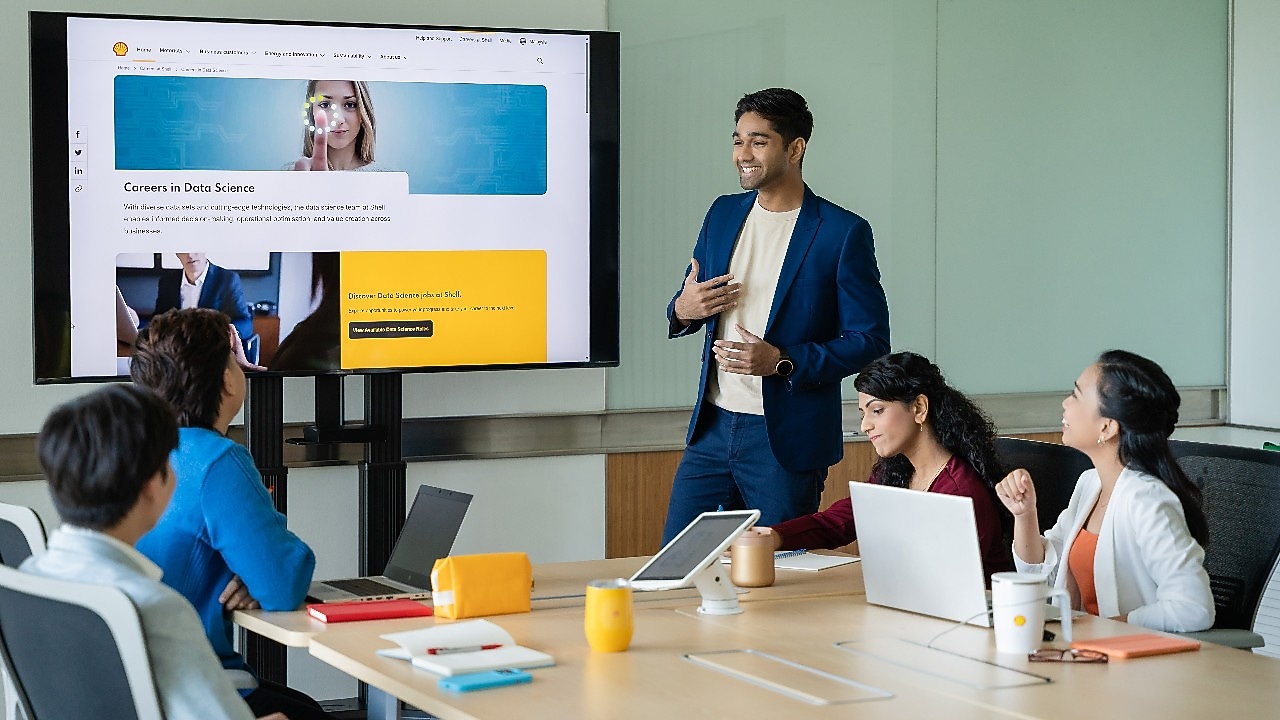 A person in a blue suit is standing and presenting to a group in a modern conference room. The presentation is displayed on a large screen showing a webpage titled “Careers in Data Science.” Four individuals are seated around a wooden meeting table with laptops, notebooks, and yellow mugs in front of them. The setting includes a clean, professional environment with glass walls and a collaborative atmosphere.​