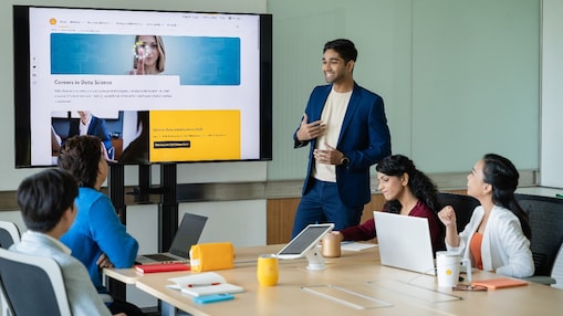 A person in a blue suit is standing and presenting to a group in a modern conference room. The presentation is displayed on a large screen showing a webpage titled “Careers in Data Science.” Four individuals are seated around a wooden meeting table with laptops, notebooks, and yellow mugs in front of them. The setting includes a clean, professional environment with glass walls and a collaborative atmosphere.