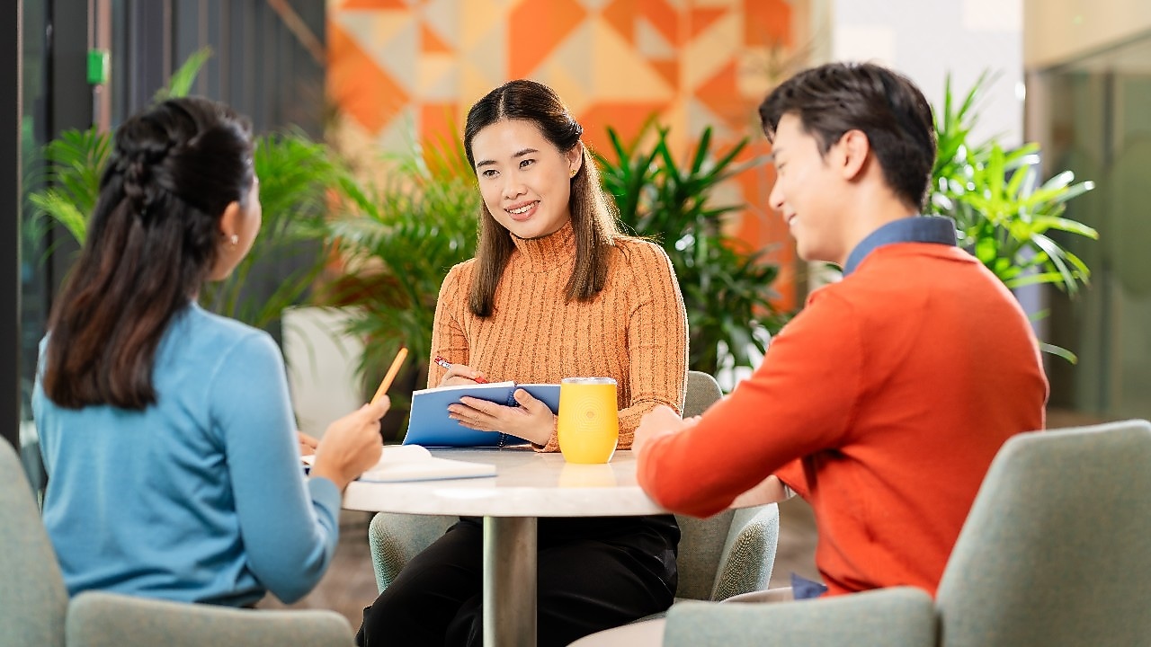 Three people are seated around a small round table in a modern office setting, engaged in a discussion. One person is holding a blue notebook, another has a yellow mug on the table, and the third appears to be gesturing while speaking. The background features green indoor plants and a wall with geometric orange patterns, creating a bright and collaborative atmosphere.