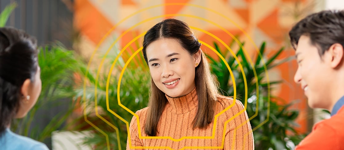 Three people sitting around a round table in a modern indoor setting, engaged in discussion. One person in an orange ribbed sweater is holding an open notebook, with a yellow mug placed on the table. The other two individuals are seated opposite, one in a blue top holding a pencil and the other in a red sweater. The background features green plants and a colorful geometric wall design.