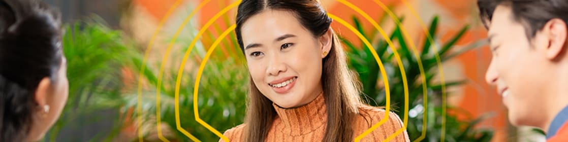 Three people sitting around a round table in a modern indoor setting, engaged in discussion. One person in an orange ribbed sweater is holding an open notebook, with a yellow mug placed on the table. The other two individuals are seated opposite, one in a blue top holding a pencil and the other in a red sweater. The background features green plants and a colorful geometric wall design.