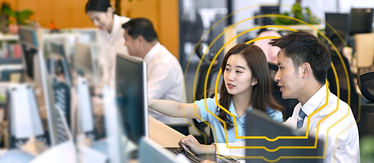 Two people sitting side by side at a desk in an open office environment, looking at a computer monitor. One person is pointing at the screen while the other observes. Multiple computer monitors and office supplies are visible on the desks, with other colleagues working in the background. The setting includes bright lighting, plants, and a modern workspace design.