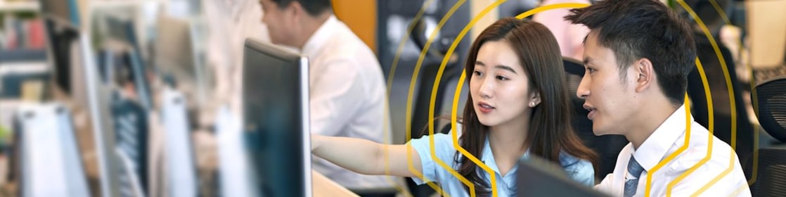 Two people sitting side by side at a desk in an open office environment, looking at a computer monitor. One person is pointing at the screen while the other observes. Multiple computer monitors and office supplies are visible on the desks, with other colleagues working in the background. The setting includes bright lighting, plants, and a modern workspace design.