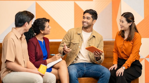 Four people are seated on a wooden bench in a modern office setting, engaged in a discussion. One person is holding an orange tablet, another has a notebook and pen, and a third person is holding a yellow mug. The background features a wall with geometric orange and beige patterns and some indoor plants, creating a vibrant and collaborative atmosphere.​