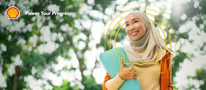 Person wearing a light beige headscarf and an orange blazer over a cream ribbed top, holding a turquoise laptop sleeve. The individual is standing outdoors with green trees and sunlight in the background, creating a bright and natural setting.