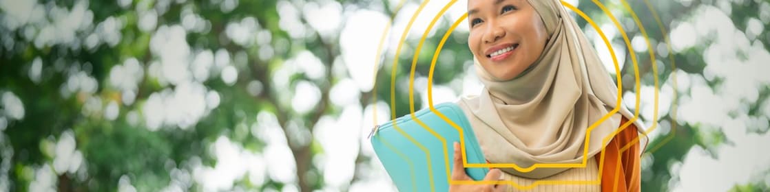 Person wearing a light beige headscarf and an orange blazer over a cream ribbed top, holding a turquoise laptop sleeve. The individual is standing outdoors with green trees and sunlight in the background, creating a bright and natural setting.