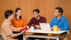 Four people are seated around a rectangular table in a meeting or discussion setting. One person on the left is holding an open red book, while another person in the center has hands resting on the table. A laptop is placed in front of the person on the right, along with two mugs—one blue and one yellow—on the table. The background features a wooden wall and two black pendant lights hanging above the group.​  ​  ​