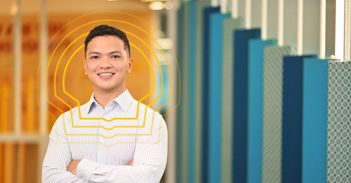 Person standing indoors with arms crossed, wearing a light blue striped button-up shirt and dark trousers. The background features a modern office setting with blue and patterned wall panels on the right and a warm-toned corridor on the left.
