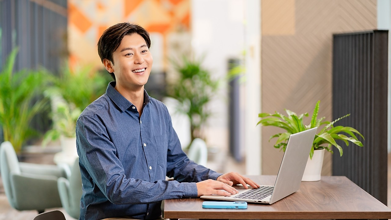 A person is seated at a wooden desk in a modern office environment, working on a laptop. A smartphone is placed on the desk next to the laptop, and a potted green plant adds a touch of decor. The background features contemporary design elements, including patterned walls, indoor plants, and lounge chairs, creating a bright and professional setting.