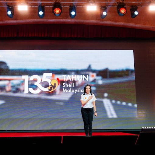 A Shell Malaysia representative speaks on stage in front of a large screen displaying the “135 Tahun Shell Malaysia” anniversary visual.