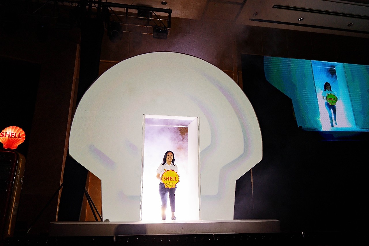 A Shell Malaysia representative makes a stage entrance through a lit Shell pecten backdrop, holding the Shell logo during the 135th anniversary media launch.