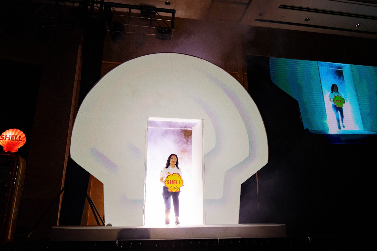 A Shell Malaysia representative makes a stage entrance through a lit Shell pecten backdrop, holding the Shell logo during the 135th anniversary media launch.