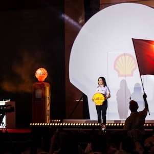 A Shell Malaysia representative stands centre stage holding the Shell logo, framed by a dramatic Shell pecten backdrop and stage lighting during the 135th anniversary launch.