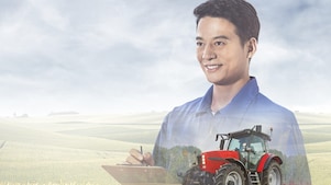 A red tractor ploughing a muddy field with trees in the background