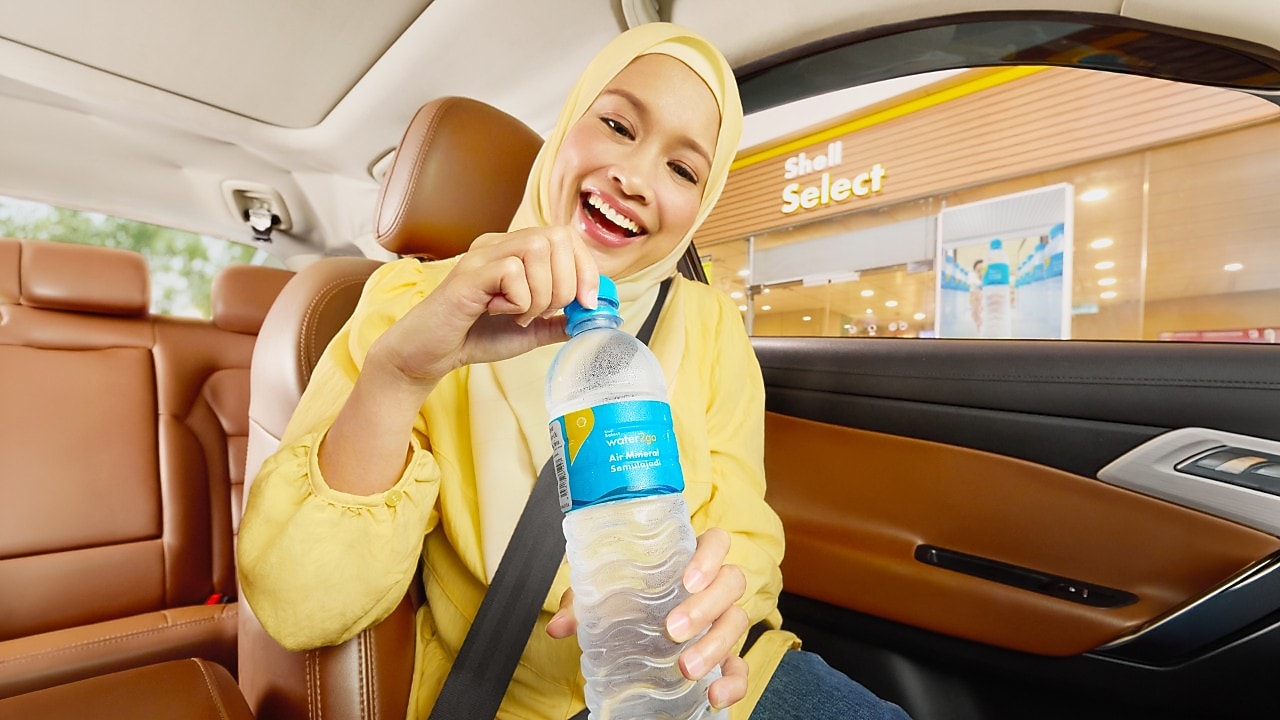 A smiling woman in a yellow outfit opens a Shell Select water2go bottle inside a car. She appears happy and relaxed, with a Shell Select store visible through the window.